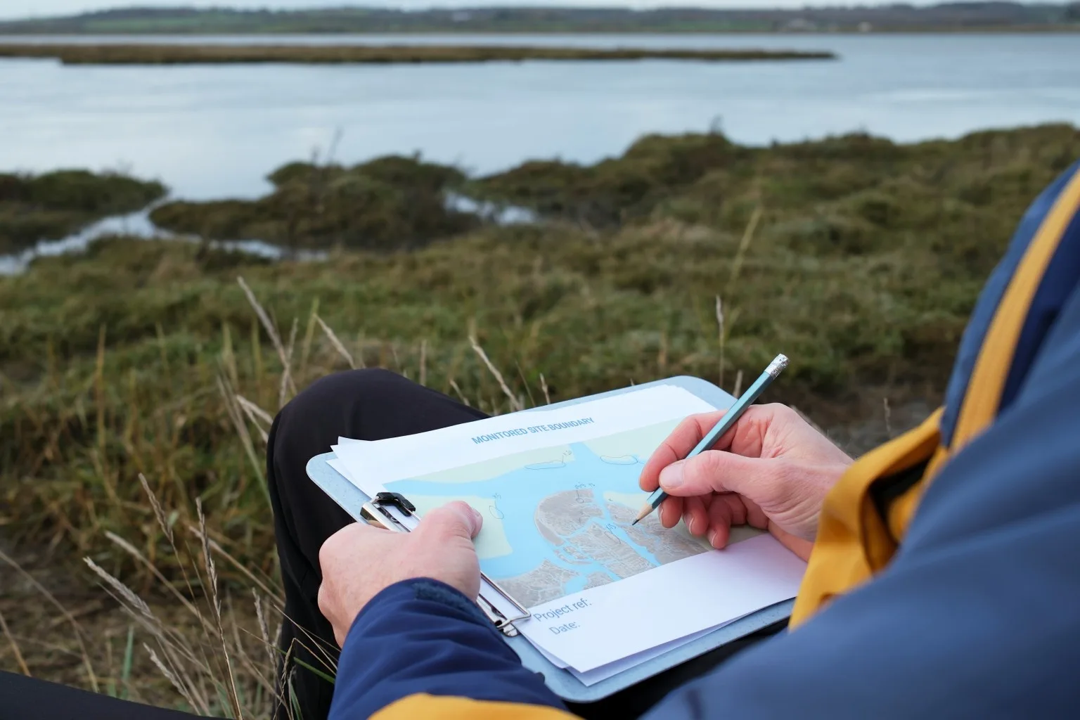 Person writing in pad of paper while sitting by lake.