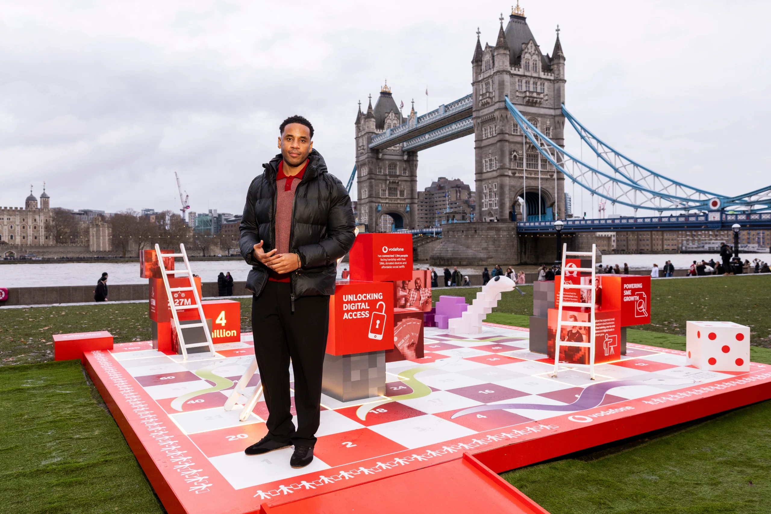 Reggie Yates standing on Vodafone UK's Snakes & Ladders installation, with Tower Bridge behind him.