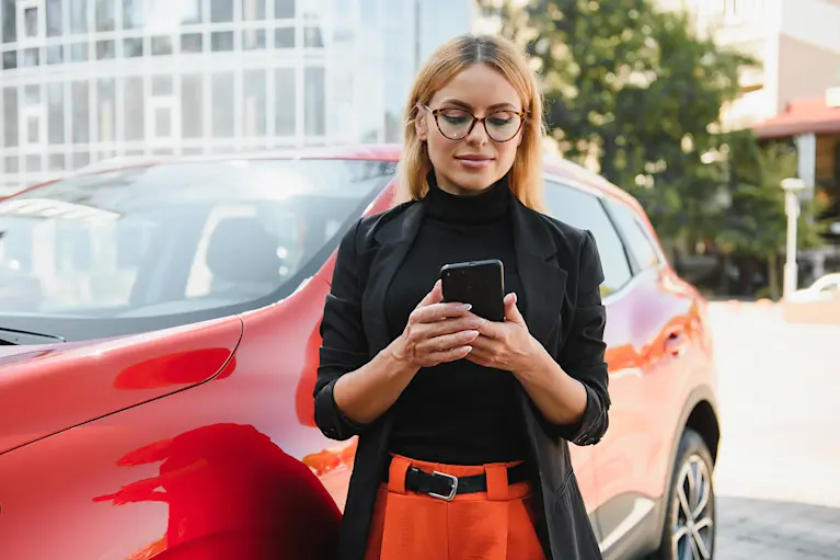 Woman using mobile phone standing by red car.