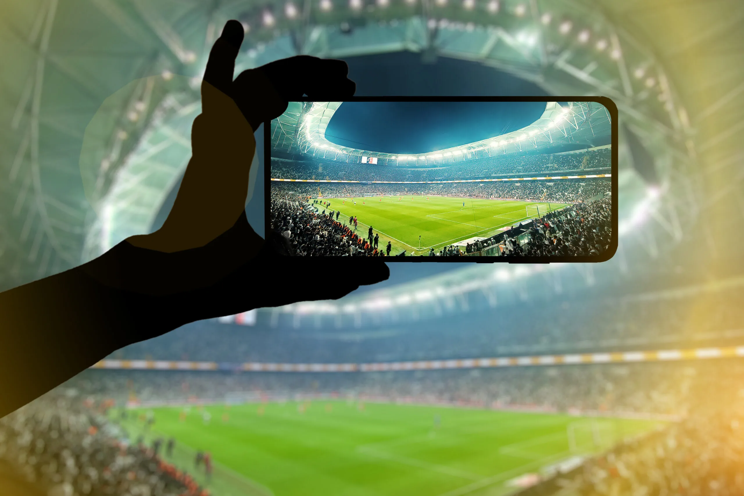 Fan hand with mobile phone photographing football game. Using a smartphone at a soccer stadium.