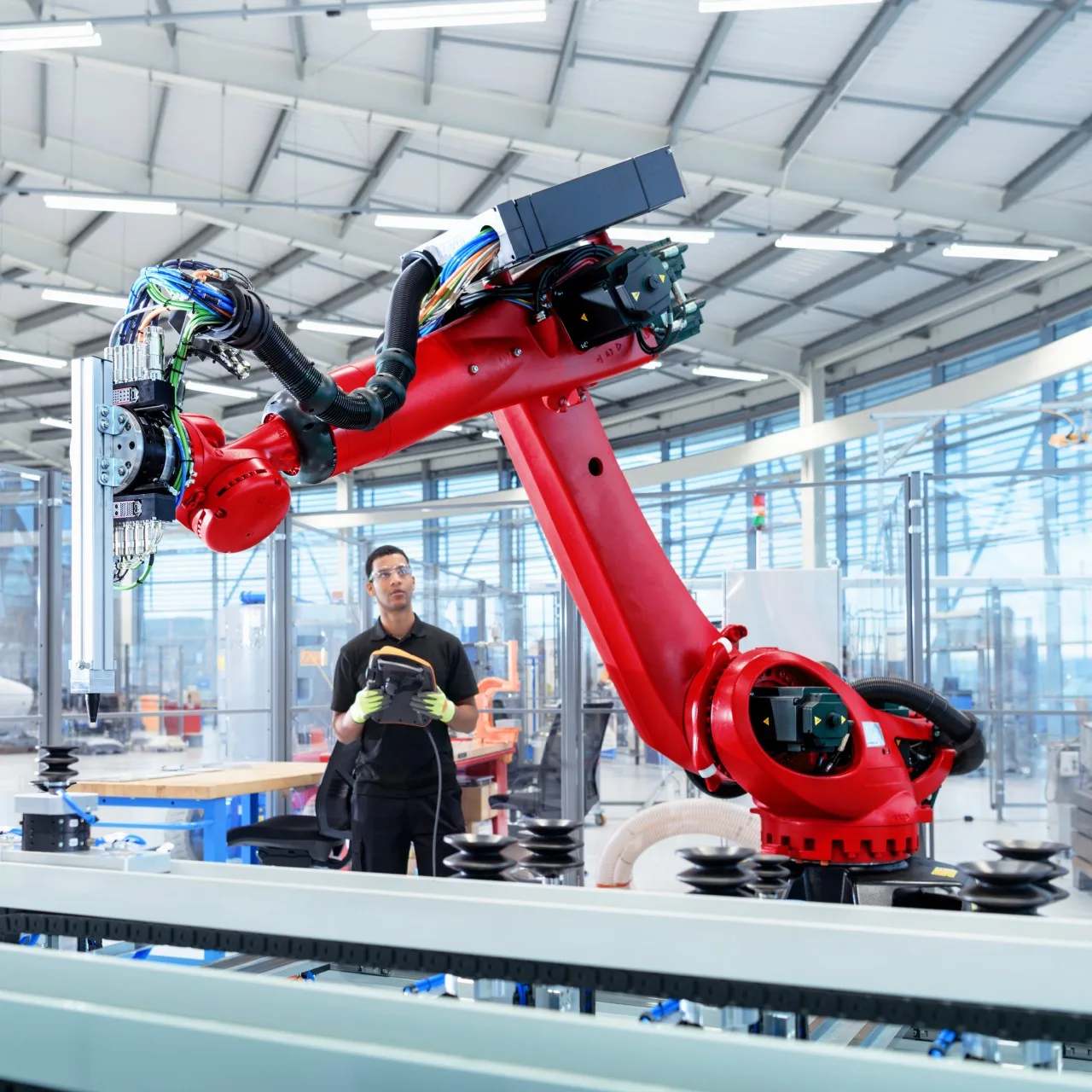 A man in safety glasses and gloves operates a large red industrial robotic arm with a control panel in a factory.