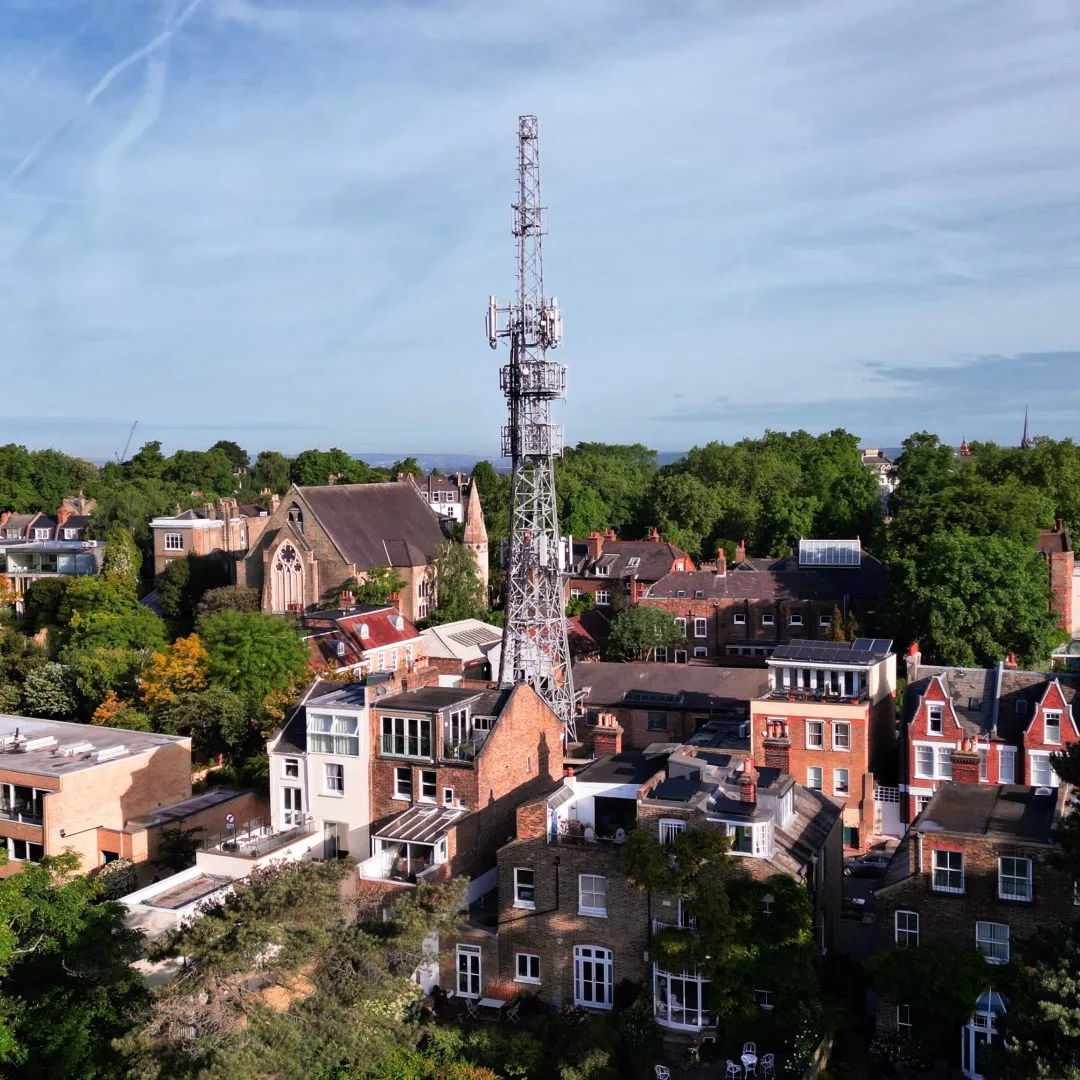 An aerial view of a tall cellular tower amidst residential buildings, a church, and abundant trees under a blue sky.