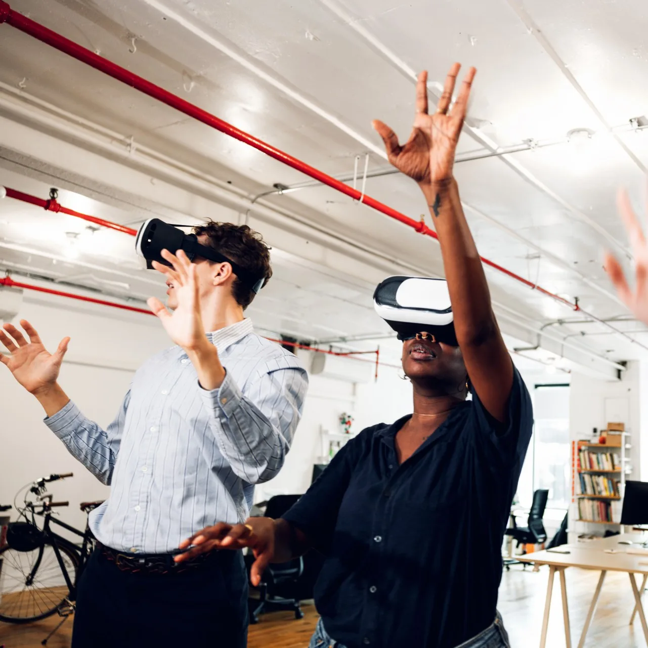 Two people wearing VR headsets, one with hands raised, in an office with exposed red pipes and a bicycle.