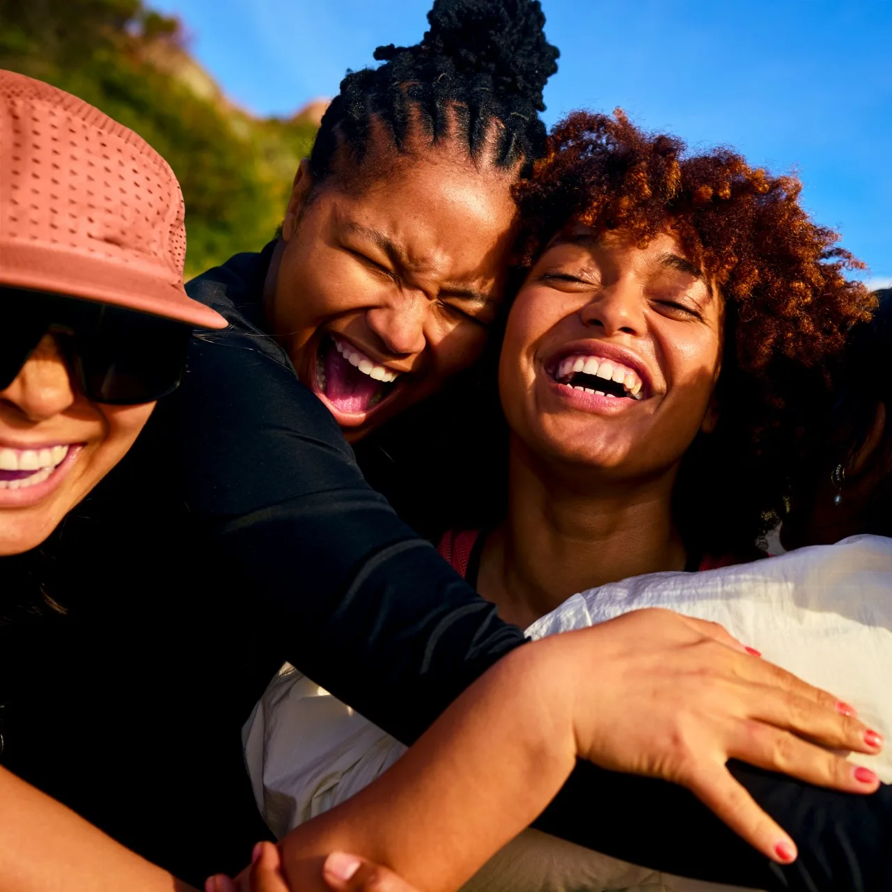 Three women embracing and laughing heartily outdoors under a blue sky. One wears a cap and sunglasses, another has braided hair, and the third has curly reddish-brown hair.