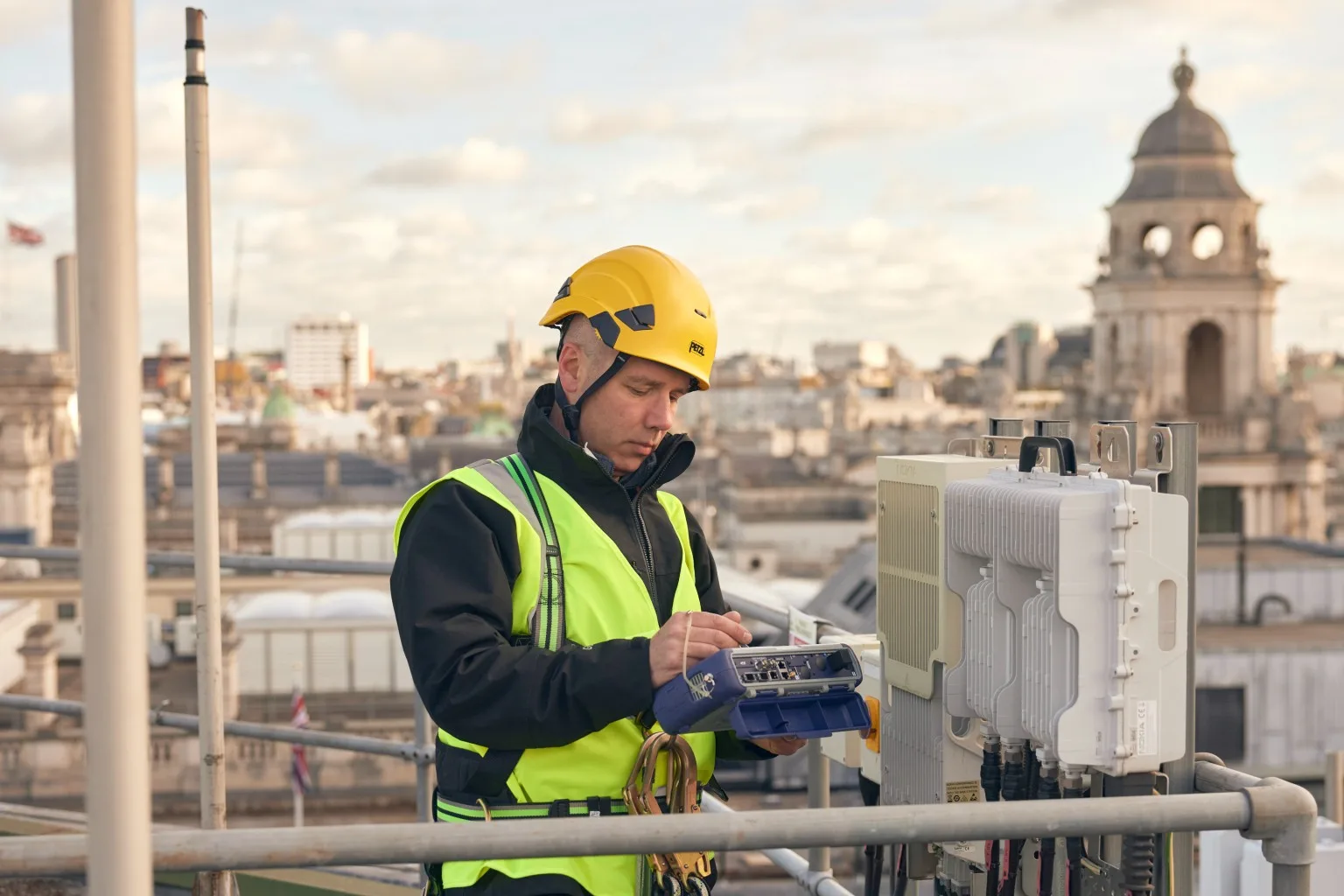 A man in a hard hat and high-visibility vest works on electronic equipment on a rooftop, with city buildings in the background.