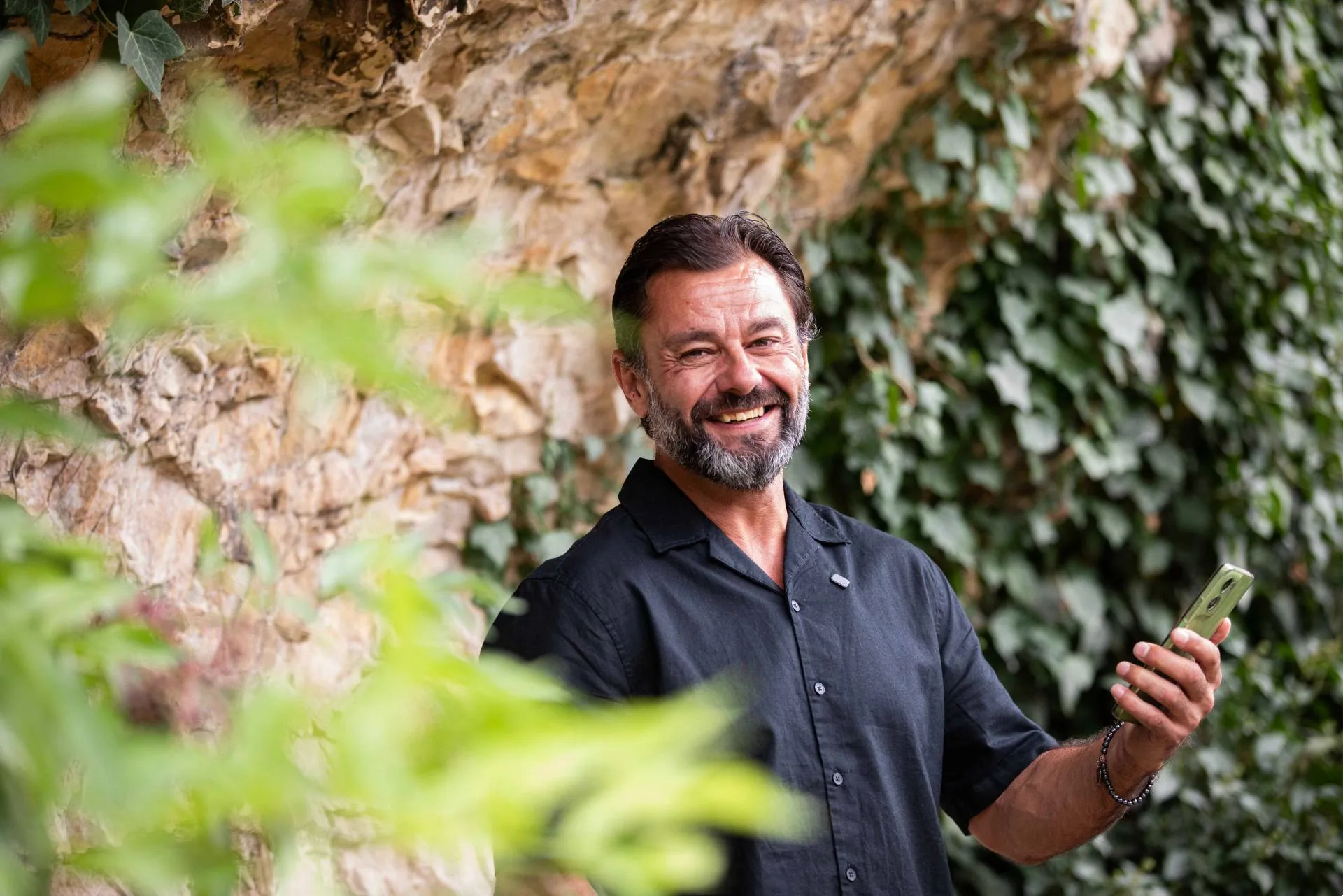 A smiling man with a beard holds a green smartphone outdoors, with a rocky wall and ivy in the background.