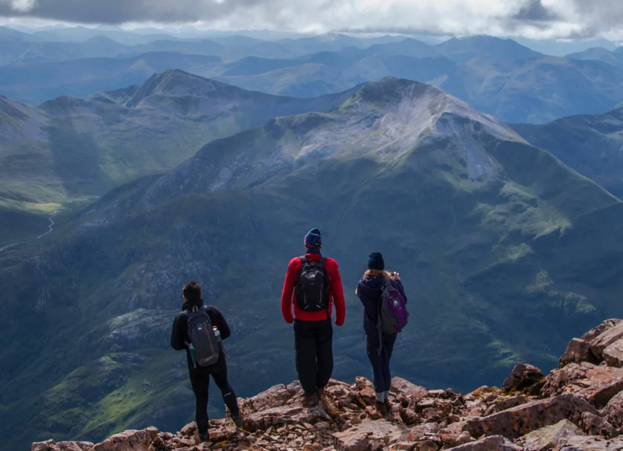 Three hikers with backpacks stand on a rocky mountain peak, looking out at a vast landscape of green and grey mountains and valleys under a cloudy sky.