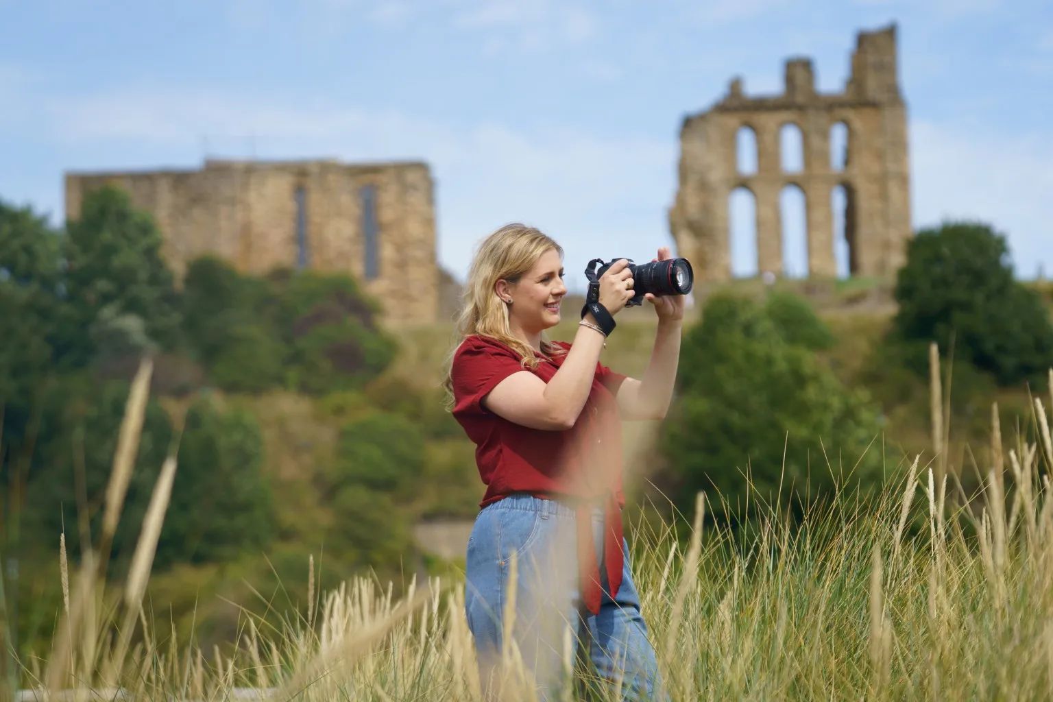 A smiling blonde woman in a red top and blue jeans holds a camera to her eye in a grassy field, with ancient stone ruins on hills in the background.