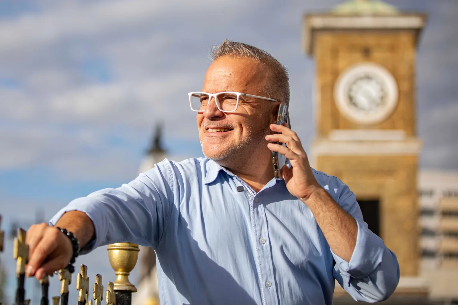 A smiling man with grey hair and white glasses talks on a phone, resting his hand on a fence, with a clock tower behind him.