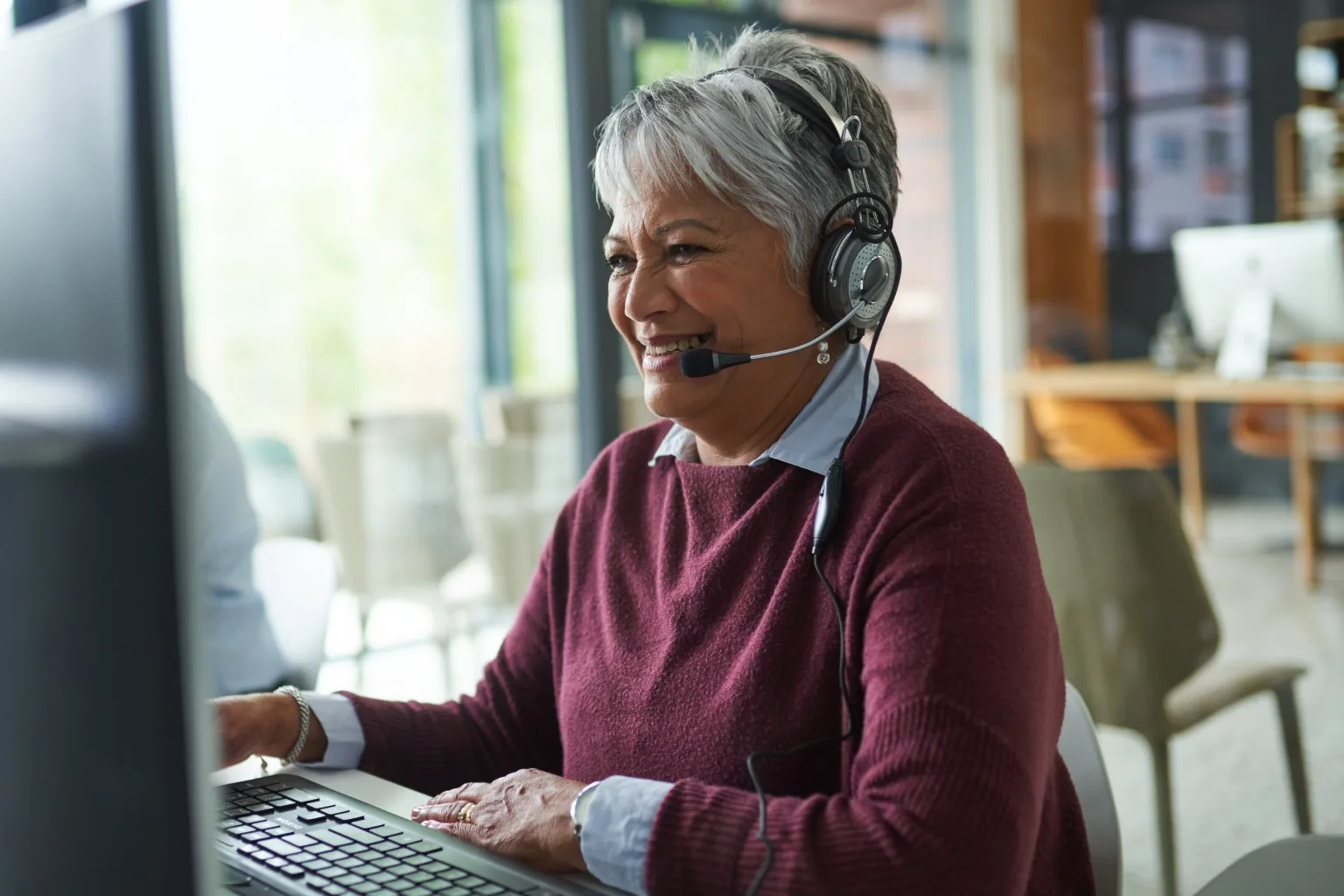 An older woman with grey hair smiles while wearing a headset and typing on a computer keyboard.