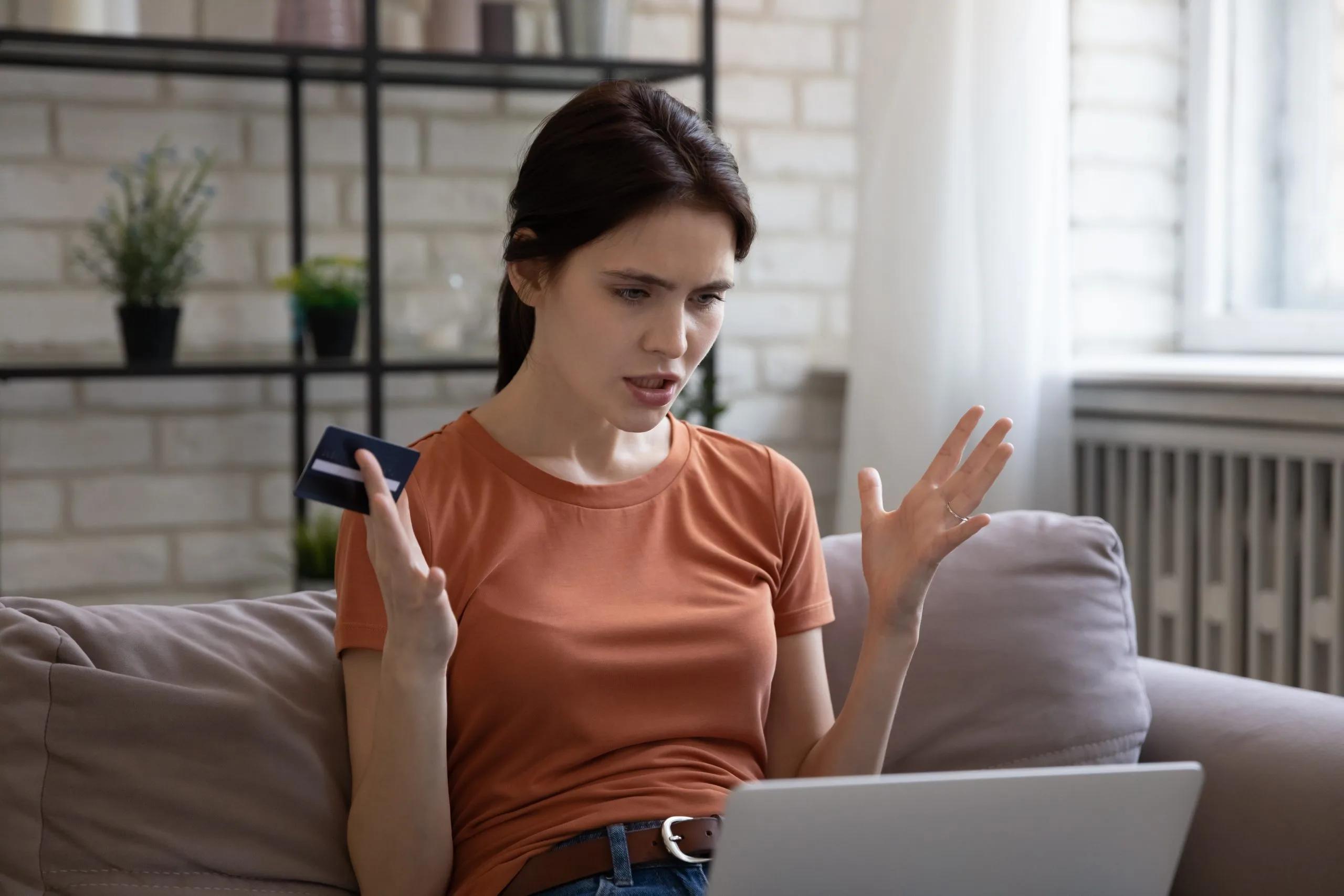 A frustrated young woman sits on a sofa, holding a credit card and gesturing at a laptop.